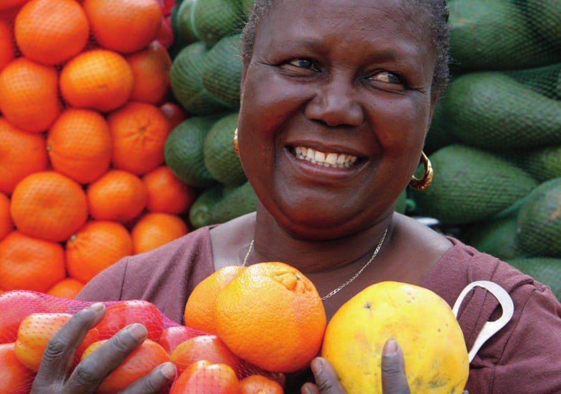 A smiling trader holds up oranges and a pawpaw in front of her fruit stall.