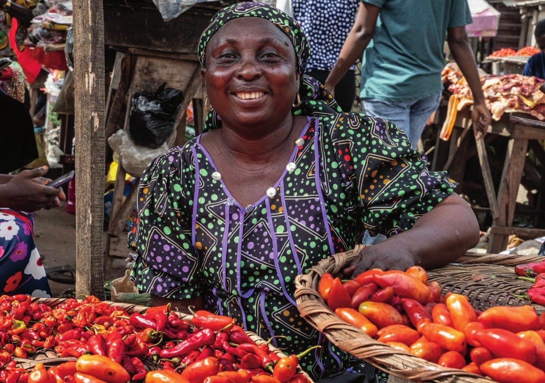 A smiling trader sits behind baskets of fresh red peppers.